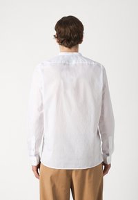 Camisa blanca de manga larga con cuello abotonado, hecha de una tela ligera y texturizada. Presenta puños abotonados y un dobladillo redondeado.