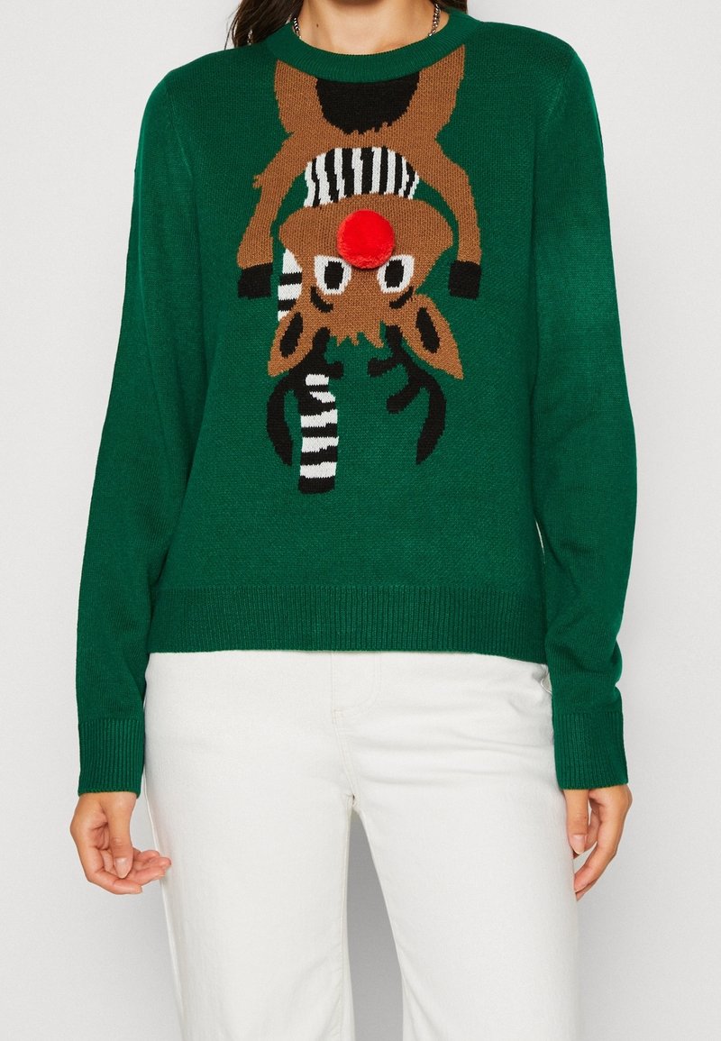 Pull vert avec un renne marron portant une écharpe rayée, avec un nez pompon rouge en 3D, porté avec un pantalon blanc.