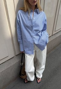 Blue and white striped button-up shirt with a relaxed fit, paired with white wide-leg trousers and black flip-flops. Brown shoulder bag visible.