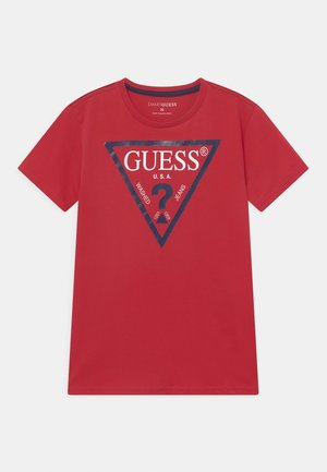 Crvena pamučna majica s trokuta grafikom. Dizajn uključuje logo i brendirane elemente "GUESS" u mornarsko plavoj boji. Kratki rukavi, okrugla izreza.
