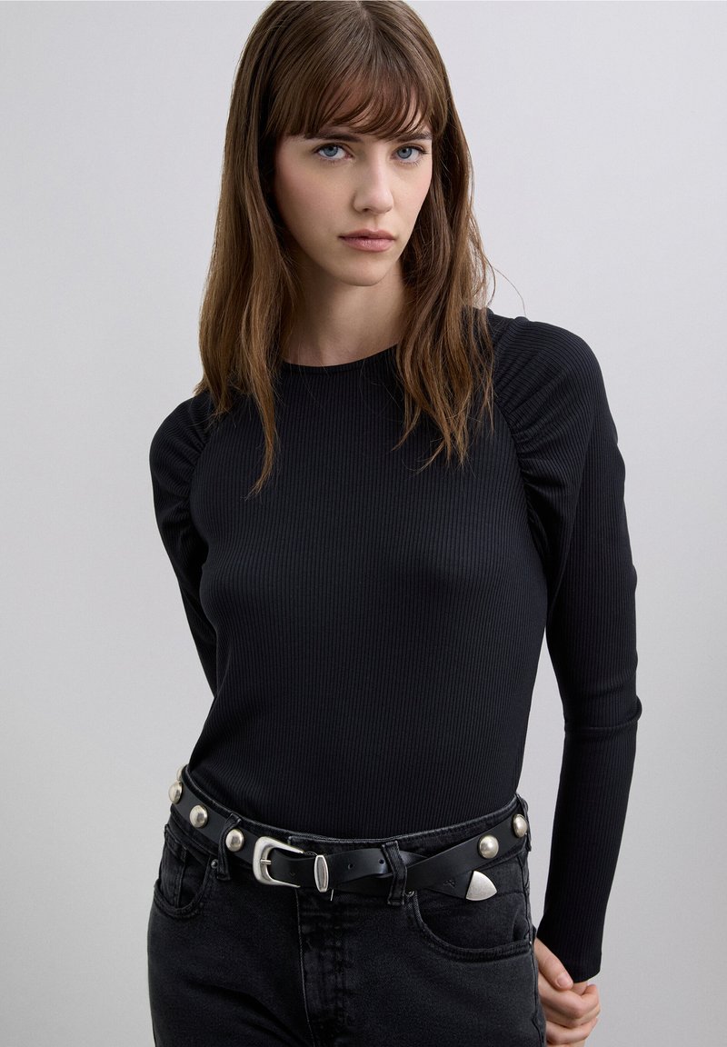 Black ribbed long-sleeve top with a rounded neckline, paired with high-waisted black jeans and a black belt featuring silver studs.