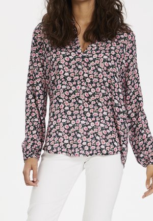 Kobieta ubrana w czarną bluzkę z długim rękawem w różowo-biały kwiatowy wzór, w połączeniu z białymi spodniami, stoi z rozluźnionymi rękami.