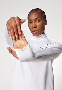 Valge tõmblukuga treeningülikond kõrge kraega, mille süles on must Nike logo. Kanga pind näeb välja sile ja liibuv.