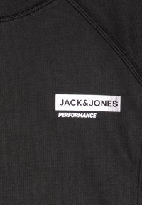 Černé sportovní tričko s texturovanou tkaninou, na kterém je umístěno bílé logo "JACK & JONES PERFORMANCE" v horním levém rohu.