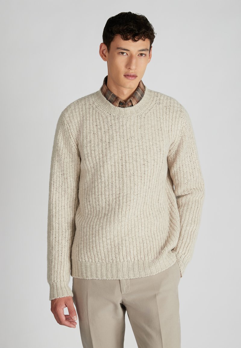 Cremfarbener, gerippter Strickpullover mit langen Ärmeln und Rundhalsausschnitt, über einem karierten Hemd getragen. Weiche Textur mit figurnaher Silhouette.