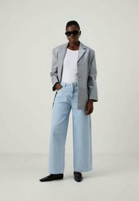 Person wearing black sunglasses, gray blazer, white shirt, light blue wide-leg jeans, and black pointed shoes, standing with one hand in pocket.