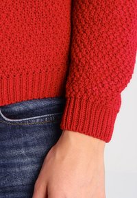 Maglione rosso a maglia con un motivo a punto testurizzato, polsini e orlo a coste. Il materiale sembra morbido, abbinato ai jeans in denim.