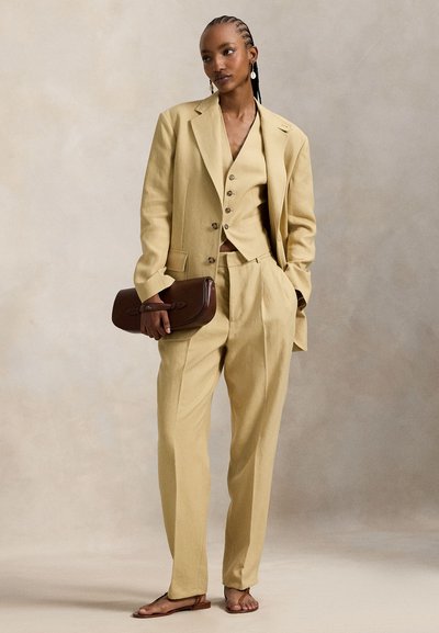 Φωτεινό κίτρινο κοστούμι τριών τεμαχίων με καλοραμμένο σακάκι, γιλέκο και παντελόνι. Το μοντέλο κρατάει ένα καφέ τσαντάκι. Ομαλό ύφασμα με ευθείες γραμμές.