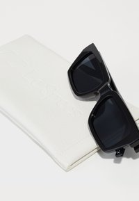 Ochelari de soare negri cu lentile întunecate, rame unghiulare, așezați pe un toc alb cu textură, gravat cu „Le Specs”. Design minimalist, fără embellishments suplimentare.