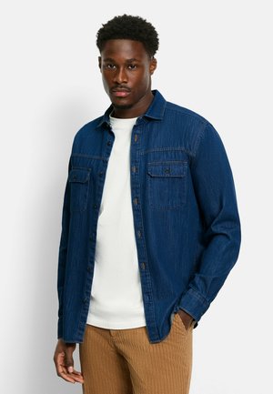 Dark blue denim shirt with a relaxed fit, front pockets, and button closures, worn over a white t-shirt, paired with brown ribbed pants.