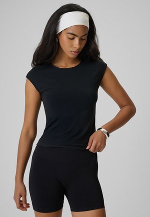 Kobieta z długimi ciemnymi włosami i białą opaską na głowie ma na sobie czarny, dopasowany t-shirt i czarne szorty, patrzy w dół z jedną ręką na biodrze.