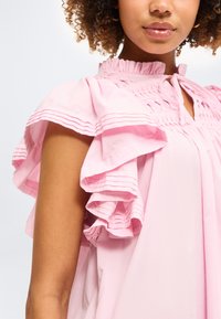 Γυναίκα με ροζ μπλούζα με στρωματοποιημένα μανίκια με βολάν και πιετέ λεπτομέρειες γύρω από το ντέκολτε και τους ώμους.