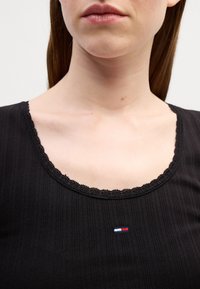 Črn rebrast top z okroglim izrezom in čipkastim robom. Ima majhen vezen logotip v rdeči, beli in modri barvi na prsih.