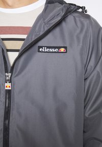 Gros plan d'une veste grise zippée avec patch logo « ellesse » et tirette assortie, portée sur une chemise rayée beige et marron.