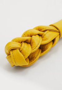 Fascetta in pelle intrecciata gialla con un design annodato, texture liscia e dettagli di cucitura a contrasto; il nodo centrale è ben evidente.