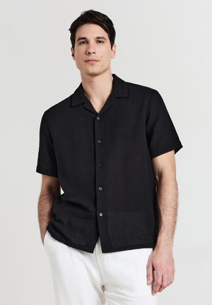 Chemise noire à manches courtes en tissu texturé ; présente un col, une fermeture à boutons et un motif horizontal subtil. Portée avec un pantalon de couleur claire.