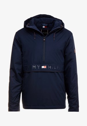 Chaqueta azul marino con capucha y cremallera frontal, texto "Tommy Hilfiger" y logo de bandera en el pecho, y parche con bandera en la manga izquierda.