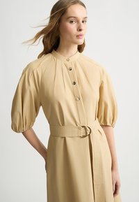 Beiges Kleid mit Puffärmeln, Stehkragen, Knopfleiste und tailliertem Gürtel. Der Stoff ist glatt mit subtiler Struktur und hat eine definierte Form.