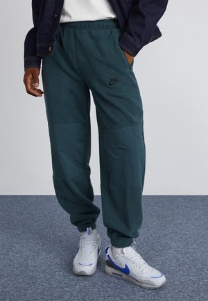 Homme portant un pantalon polaire Nike vert foncé et des baskets blanches Nike avec des virgules bleues, debout sur un tapis gris.