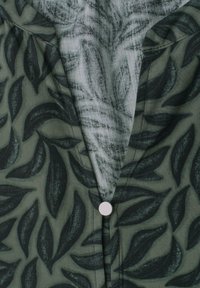 Tissu vert foncé avec motif de feuilles noires, doublure intérieure claire et un seul bouton rond blanc fermant le vêtement.