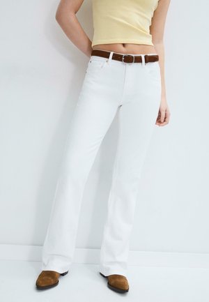 Kobieta ubrana w białe dżinsy z wysokim stanem i rozszerzanymi nogawkami, brązowy pasek, żółty krótki top oraz brązowe zamszowe buty na szpic, stojąca przy białej ścianie.