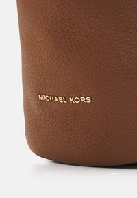 Barna bőr pehely kézitáska közeli nézete, arany színű "MICHAEL KORS" logóval. Texturált felület, lekerekített aljjal és varrott részletekkel.