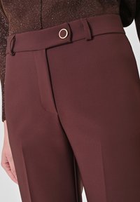 Pantaloni eleganti color bordeaux con una texture liscia, caratterizzati da un bottone circolare color oro e una vestibilità su misura. Design a vita piatta con cuciture sottili.