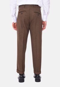 Pantaloni in lana marrone con vestibilità affusolata, due tasche posteriori con bottoni e orli a pieghe. Indossati con una camicia bianca a maniche lunghe.