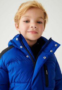 Zils pūkains jaka ar ieņemamu fronti, kam ir aizdare ar rāvējslēdzēju, spiedpogu aizdari un melniem akcentiem uz pleciem. Iekļauj kapuci un quilting tekstūru.