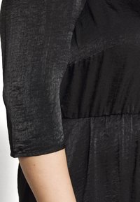 Manche de robe en tissu noir texturé se terminant juste en dessous du coude sur une personne avec un avant-bras et une main visibles, sur un fond blanc.