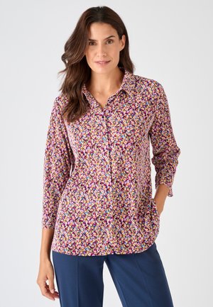 Femme portant un chemisier à manches longues avec un motif floral multicolore, associé à un pantalon bleu marine, debout avec une main dans la poche.