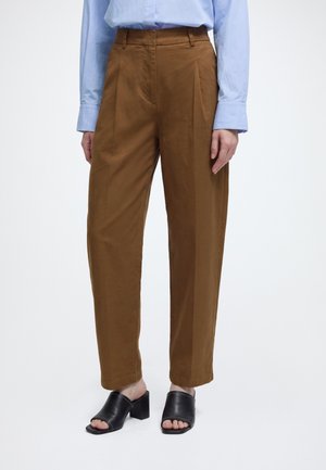 Personne portant un pantalon marron taille haute à jambes droites, une chemise bleu clair à manches longues et des sandales noires ouvertes à talons, debout devant un fond uni.