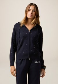 Sweat à zip bleu marine avec des embellissements en bijoux, manches longues et capuche avec cordon, associé à un pantalon bleu marine assorti avec un logo décoratif.