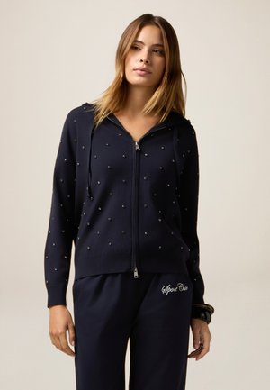 Sweat à zip bleu marine avec des embellissements en bijoux, manches longues et capuche avec cordon, associé à un pantalon bleu marine assorti avec un logo décoratif.