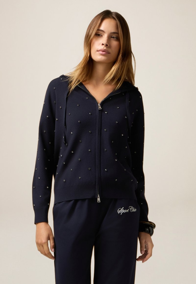 Sweat à zip bleu marine avec des embellissements en bijoux, manches longues et capuche avec cordon, associé à un pantalon bleu marine assorti avec un logo décoratif.