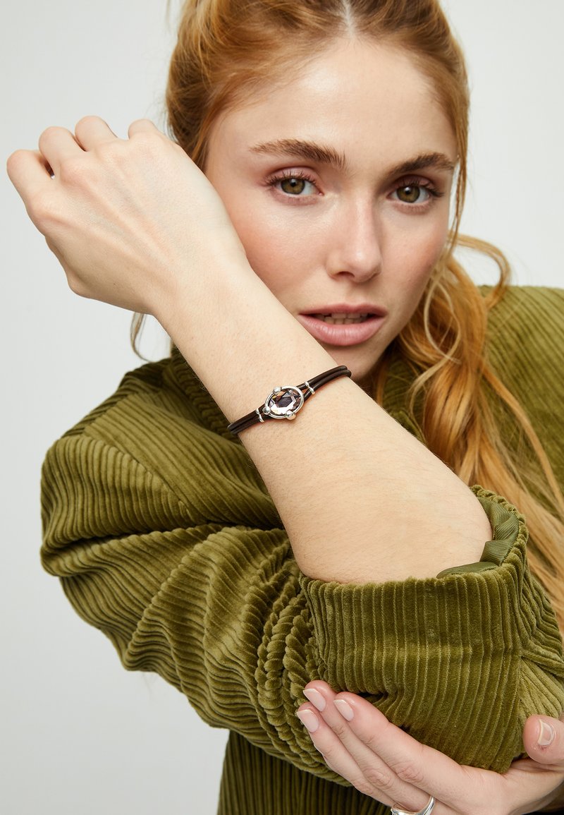 Bracelet en cuir avec un accent en argent orné d'une pierre violette, porté au poignet, sur un vêtement vert olive texturé.