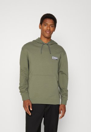 Sudadera verde oliva con cordón, bolsillo tipo canguro y pequeño parche gráfico en el pecho. Hecha de una tela suave y texturada.