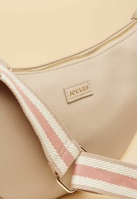 Beige strukturierte Umhängetasche mit goldenem Reißverschluss, verstellbarem Träger mit rosa-weißen Streifen und goldenem "JOULES"-Logo-Patch.