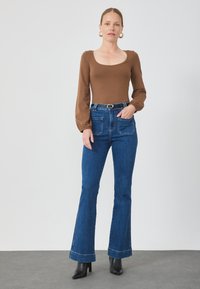 Brauner Langarm-Body mit quadratischem Ausschnitt, kombiniert mit hoch taillierten blauen Schlagjeans. Schwarze Absatz-Stiefeletten runden das Outfit ab.