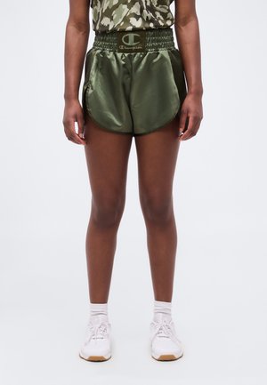 Άτομο που φοράει πράσινα σατέν αθλητικά σορτς με ελαστική μέση και λευκά αθλητικά παπούτσια, στέκεται μπροστά σε ένα απλό ανοιχτό φόντο.