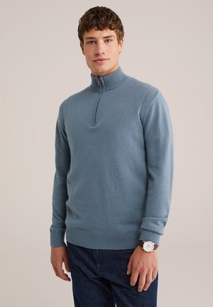 Pull-over bleu clair à zip, fabriqué en tissu doux, avec des manches longues et des poignets côtelés. Porté avec un jean foncé et une montre-bracelet.