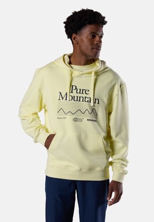 Mann trägt hellgelben Hoodie mit Aufschrift "Pure Mountain" und Berggrafik, kombiniert mit dunkelblauer Hose, steht mit einer Hand in der Tasche.