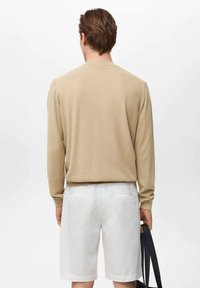 Beige long-sleeve knit sweater paired with white shorts, held with a dark bag. Textured fabric, simple design, visible seam details.