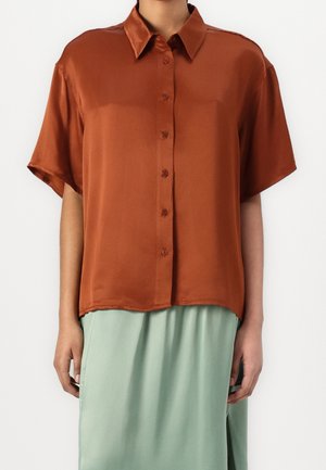 Vrouw draagt een roestkleurige satijnen blouse met korte mouwen en knopen, gecombineerd met een lichtgroene rok met een zijschuif.