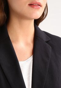 Czarna marynarka z wyraźnie zarysowanym klapem, widoczna faktura tkaniny. Noszona na jasnoszarym topie, podkreślająca okolice kołnierzyka i dekoltu.