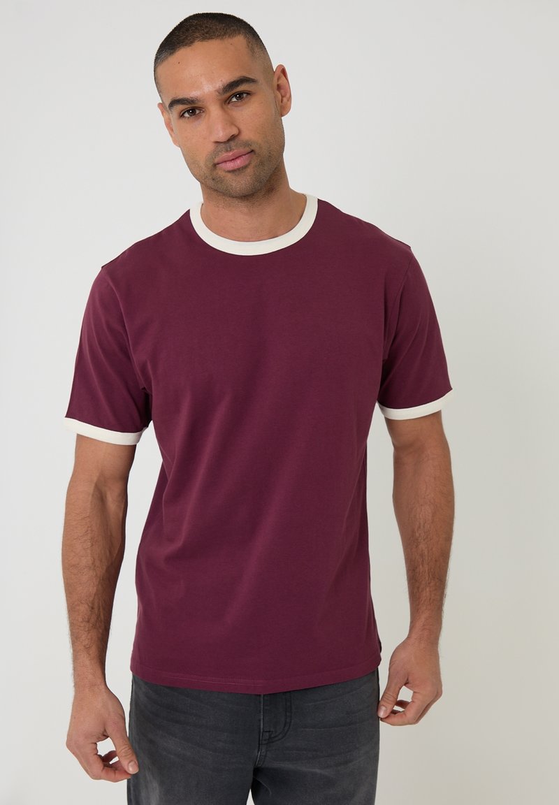 Mand med kort hår iført en bordeaux T-shirt med hvid kant ved krave og ærmer, stående mod en ensartet lys baggrund.