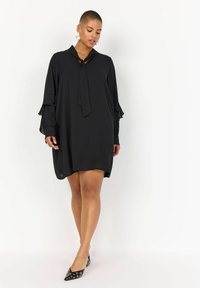 Rochie neagră cu croi lejer, mâneci lungi cu detalii din volă, și un decolteu legat. Textură de stofă netedă, lungime deasupra genunchiului.