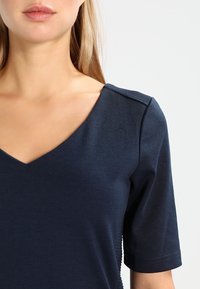 Mörkblå V-ringad topp med korta ärmar, tillverkad av slätt, stretchigt material. Har subtila ribbade detaljer vid axelsömmen.