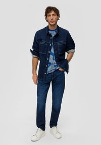 Granatowa kurtka dżinsowa na guzik, z dwiema kieszeniami na piersiach i podwiniętymi rękawami, noszona na niebieskiej koszuli w wzory, zestawiona z ciemnymi dżinsami i białymi sneakersami.