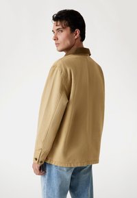 Man met donker haar in een beige jas en lichtblauwe jeans, zijwaarts gekeerd tegen een eenvoudige witte achtergrond.